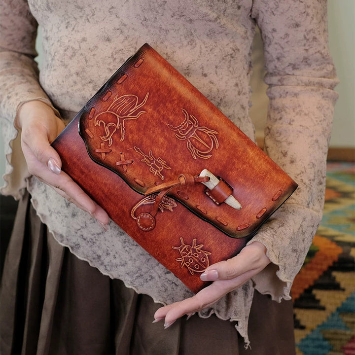 Person holding a brown A5 leather journal with hand-tooled beetle motifs.