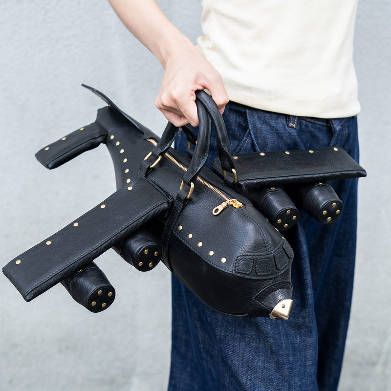 Black airplane-shaped leather handbag with gold studs, held by hand