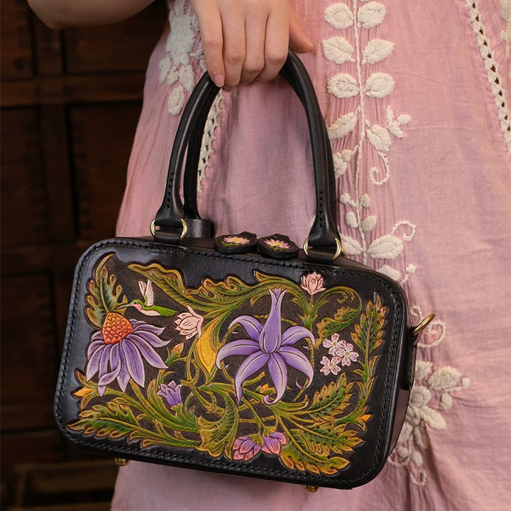 A woman holding a black floral tooled leather top handle bag with colorful flowers and a hummingbird.