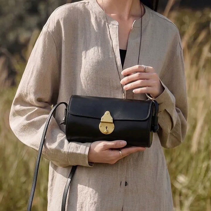 A woman in a linen shirt holds a black vegetable tanned leather barrel bag with a brass latch.