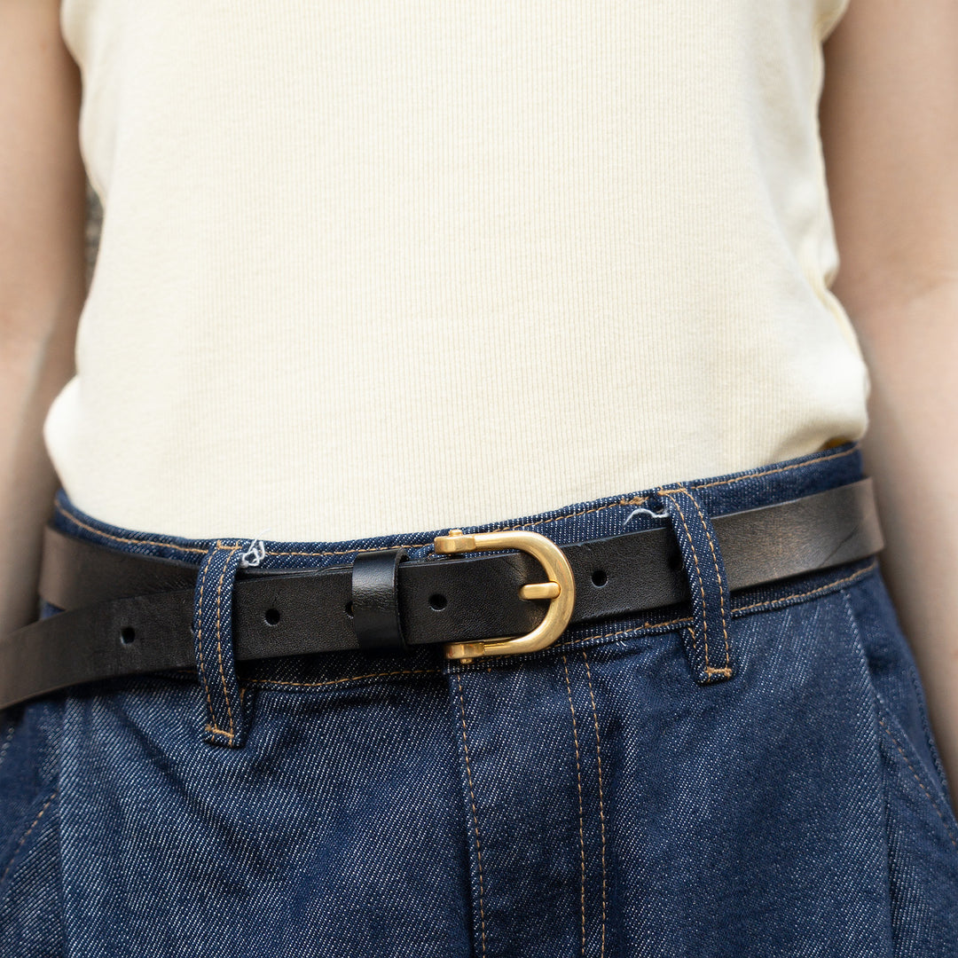 Woman wearing a slim black leather belt with a gold-tone buckle paired with blue jeans.
