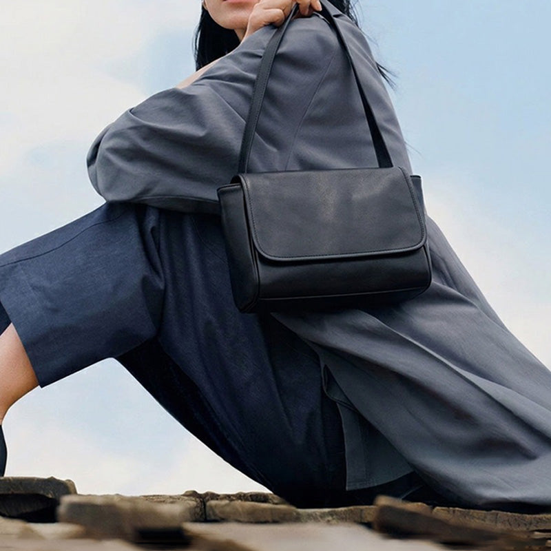 Model wearing a black leather shoulder bag with a rounded flap and a minimalist design.