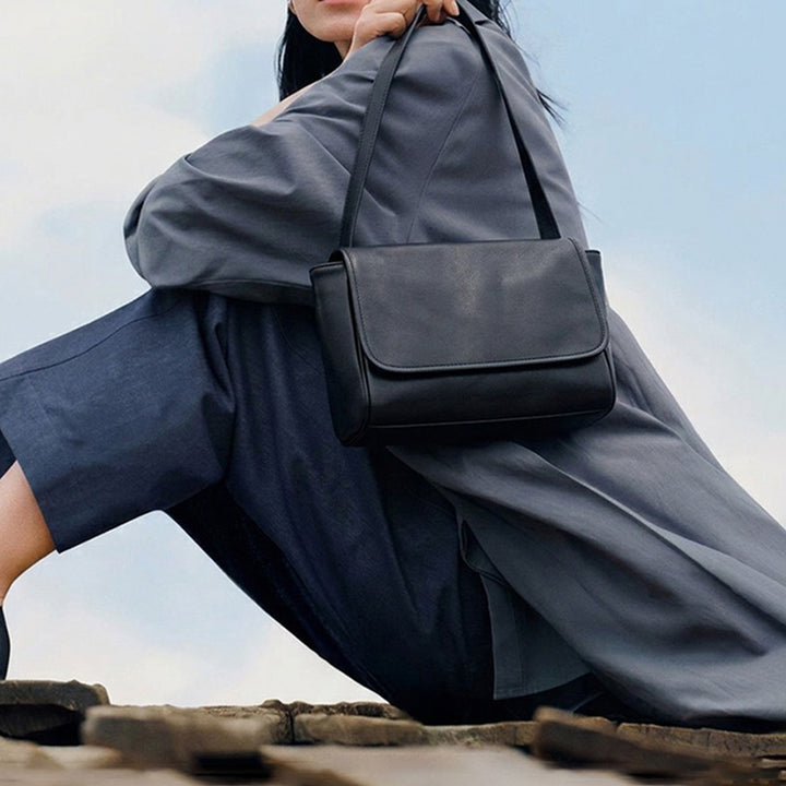 Model wearing a black leather shoulder bag with a rounded flap and a minimalist design.