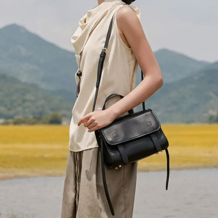 Model wearing a black leather satchel bag with a top handle and long shoulder strap.