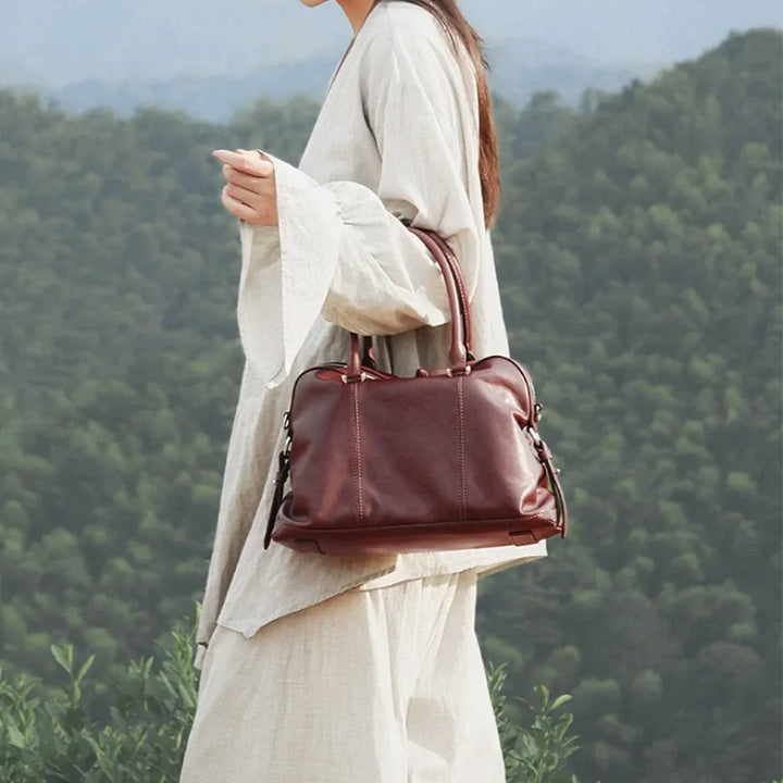 Woman carrying a brown leather Boston bag by its rounded top handles outdoors.