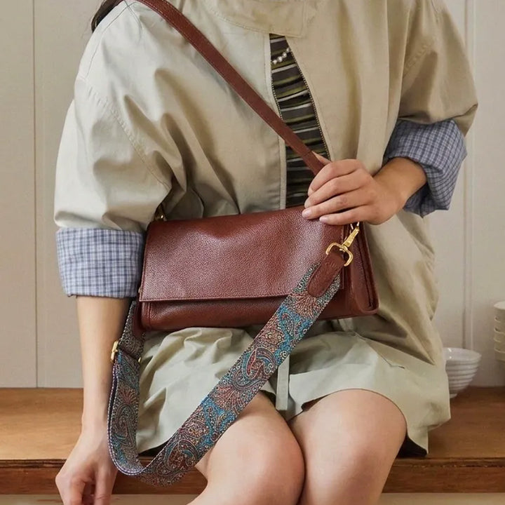 Model wearing a brown leather flap crossbody bag with a patterned strap draped beside it.