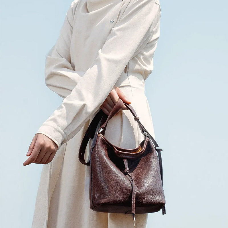 Model in a cream dress holding a brown leather hobo bag with a knotted strap and metal ring detail.