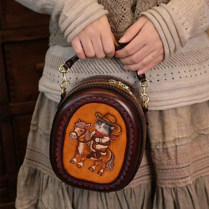 Model holding an oval leather crossbody bag with a cowboy cat riding a horse on the front.