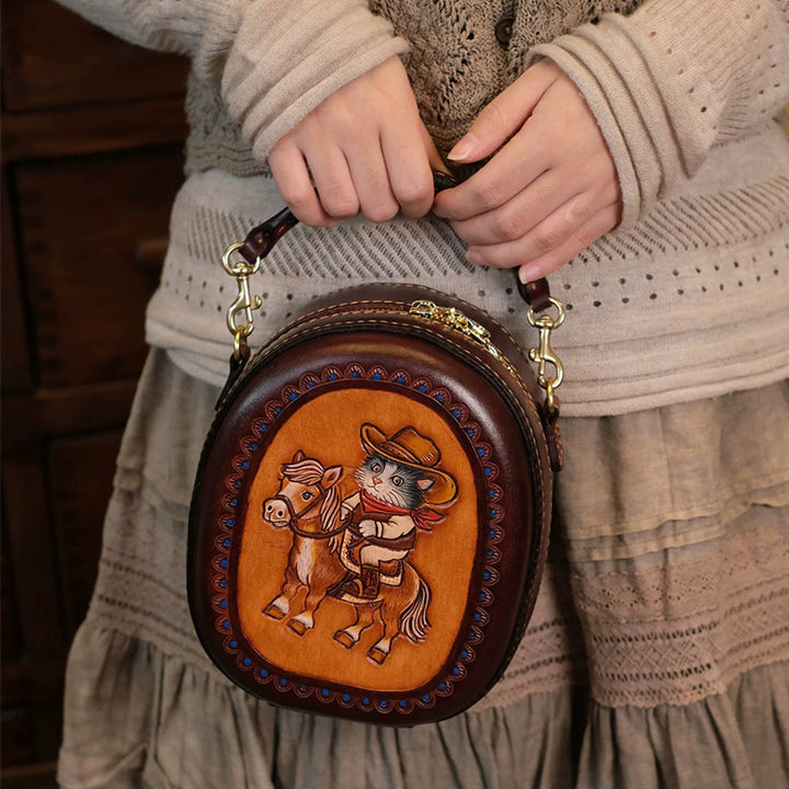 Model holding an oval leather crossbody bag with a cowboy cat riding a horse on the front.