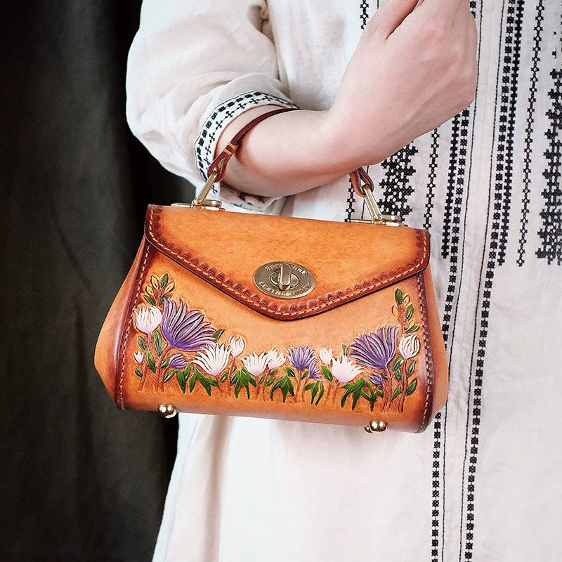 Model carrying a brown floral leather top handle bag on her forearm, showing its turn-lock flap.