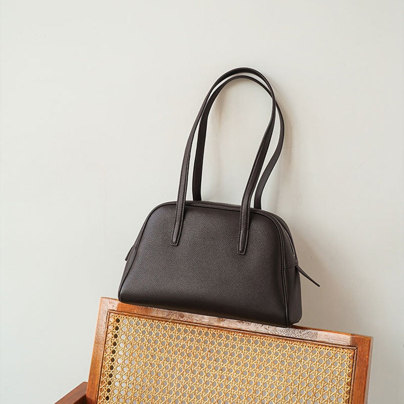 Minimalist chocolate brown leather bowling bag with twin straps resting on a rattan chair