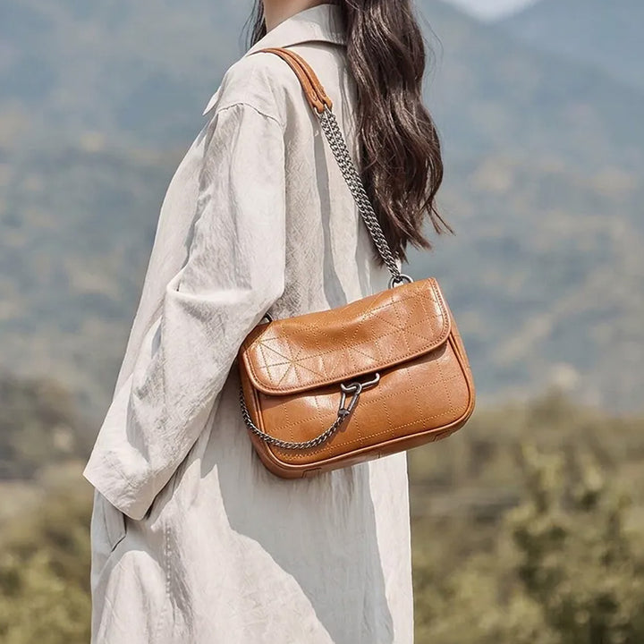 Model wearing a tan leather flap shoulder bag with a chain strap outdoors.