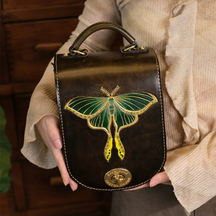 A person holds a brown leather satchel backpack with a green luna moth motif and top handle.