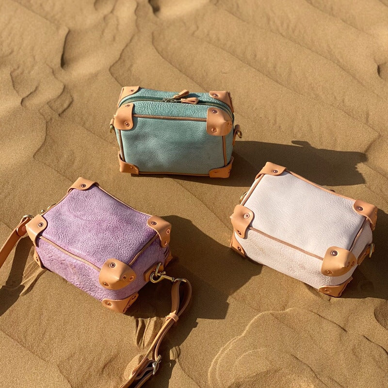Three mini trunk crossbody bags in veg tan leather laid on sand — purple, green, and apricot