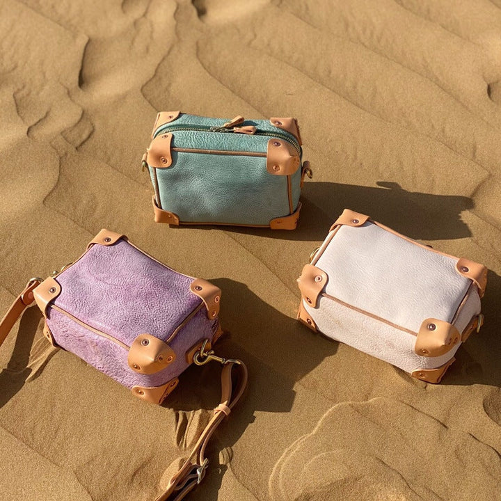 Three mini trunk crossbody bags in veg tan leather laid on sand — purple, green, and apricot