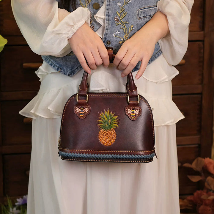 Person holding a brown leather dome bag with a pineapple motif and bee details.