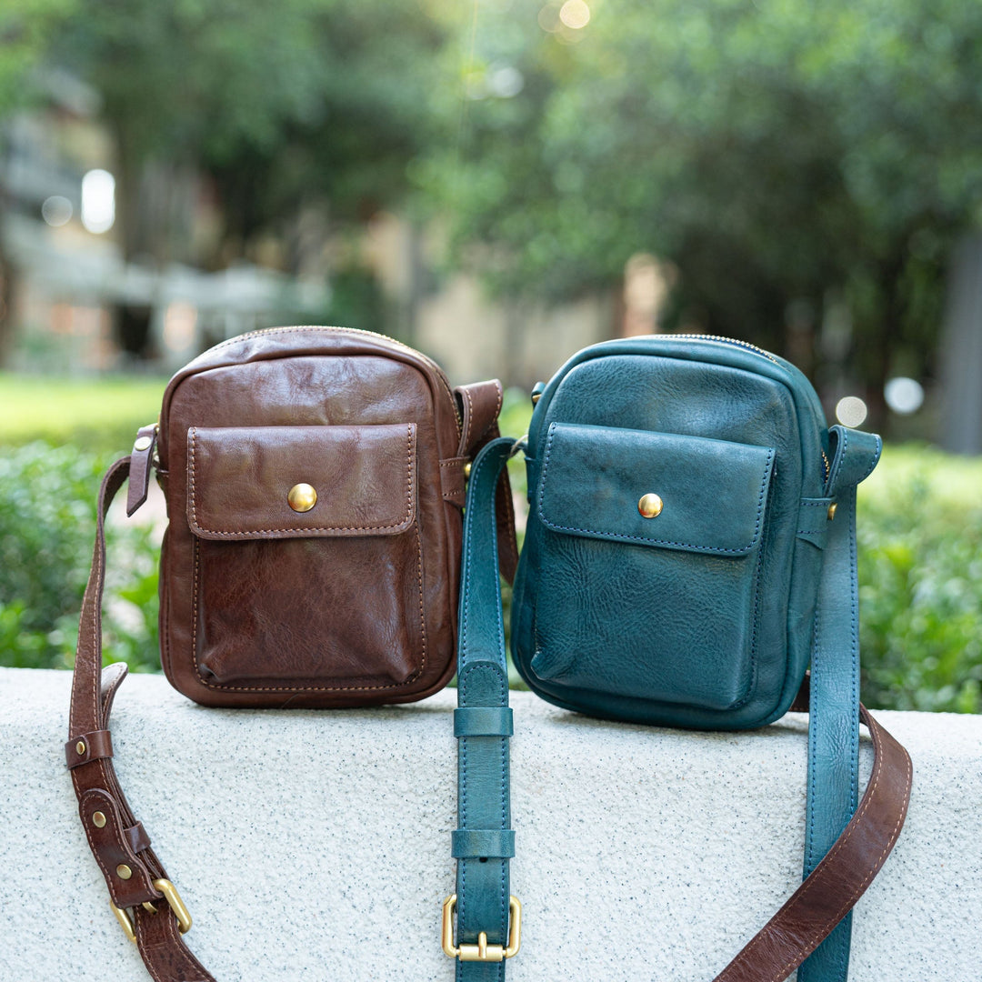 Brown and teal small leather crossbody bags with front snap pocket on stone ledge