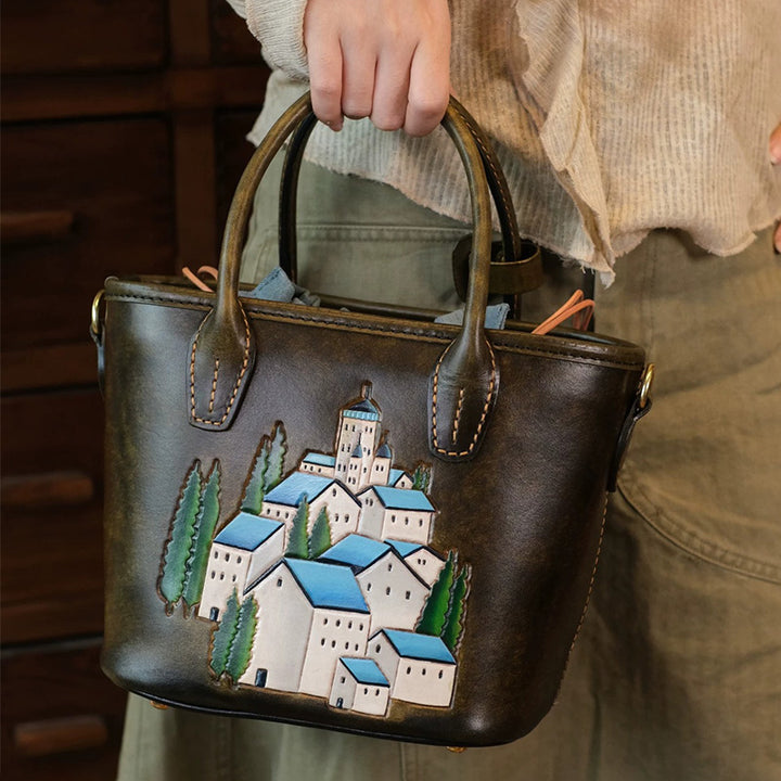 Woman holding an olive-brown leather tote with hand-tooled village artwork.