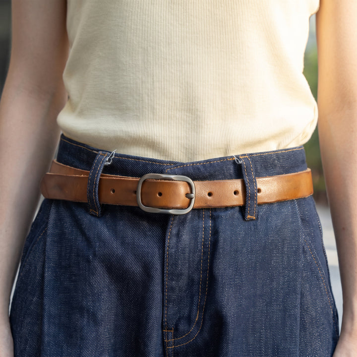 Woman wearing a slim brown leather belt with a metal pin buckle and blue jeans.